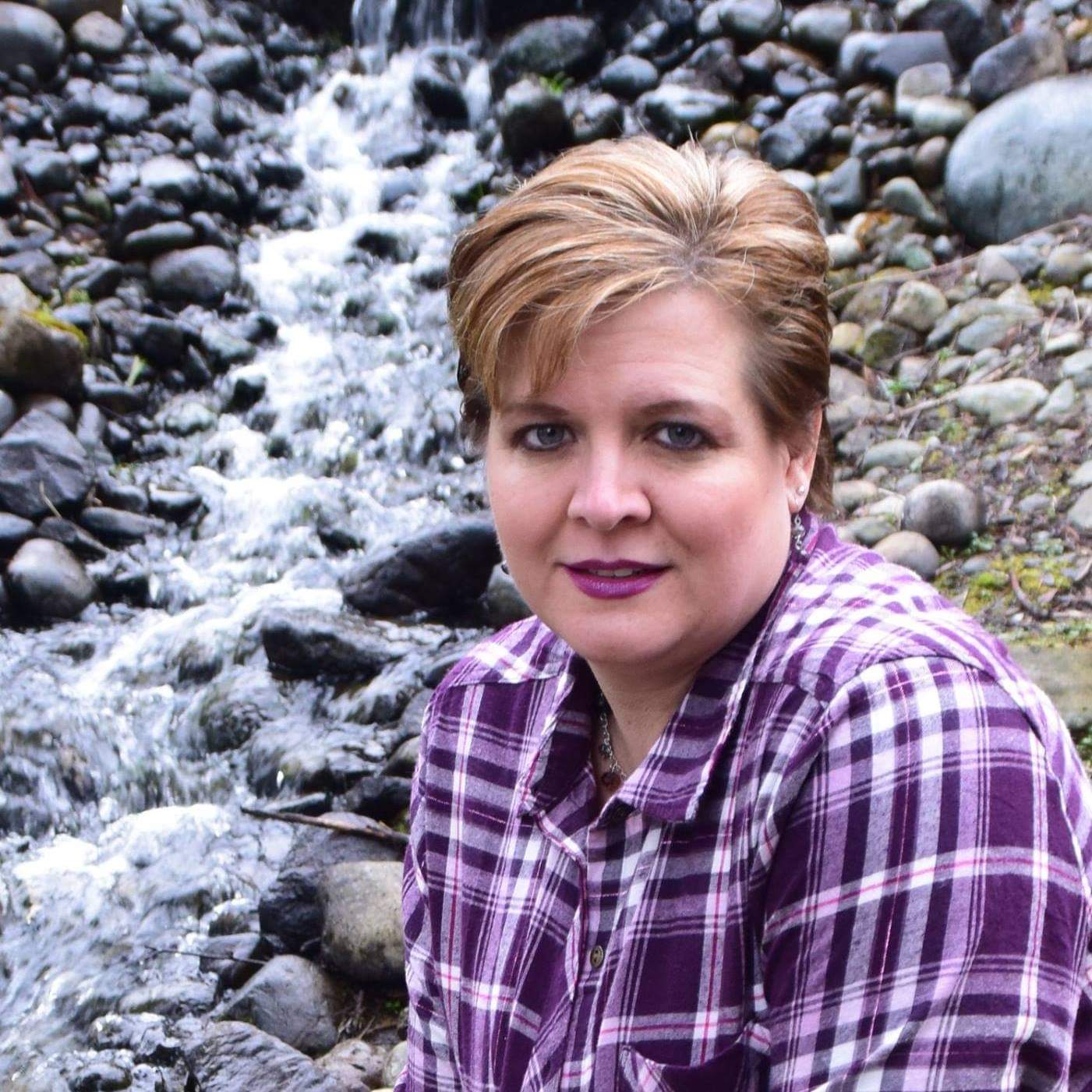 Carli, white woman with short blonde and brown hair, wearing a purple and white flannel button up top with a stream and rocks behind her