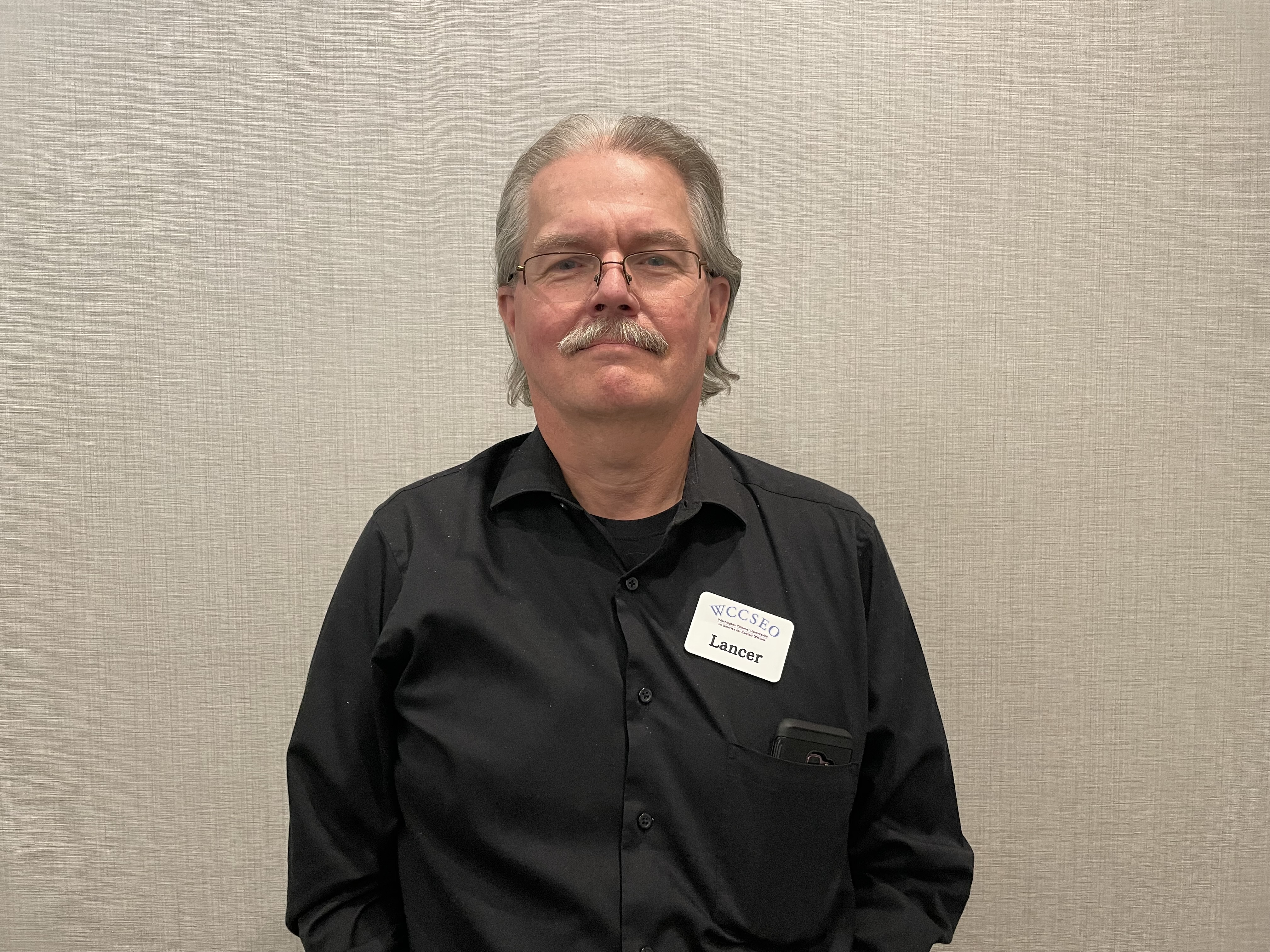 Lancer, white man with a slight smile, glasses, and a moustache, wearing a black shirt