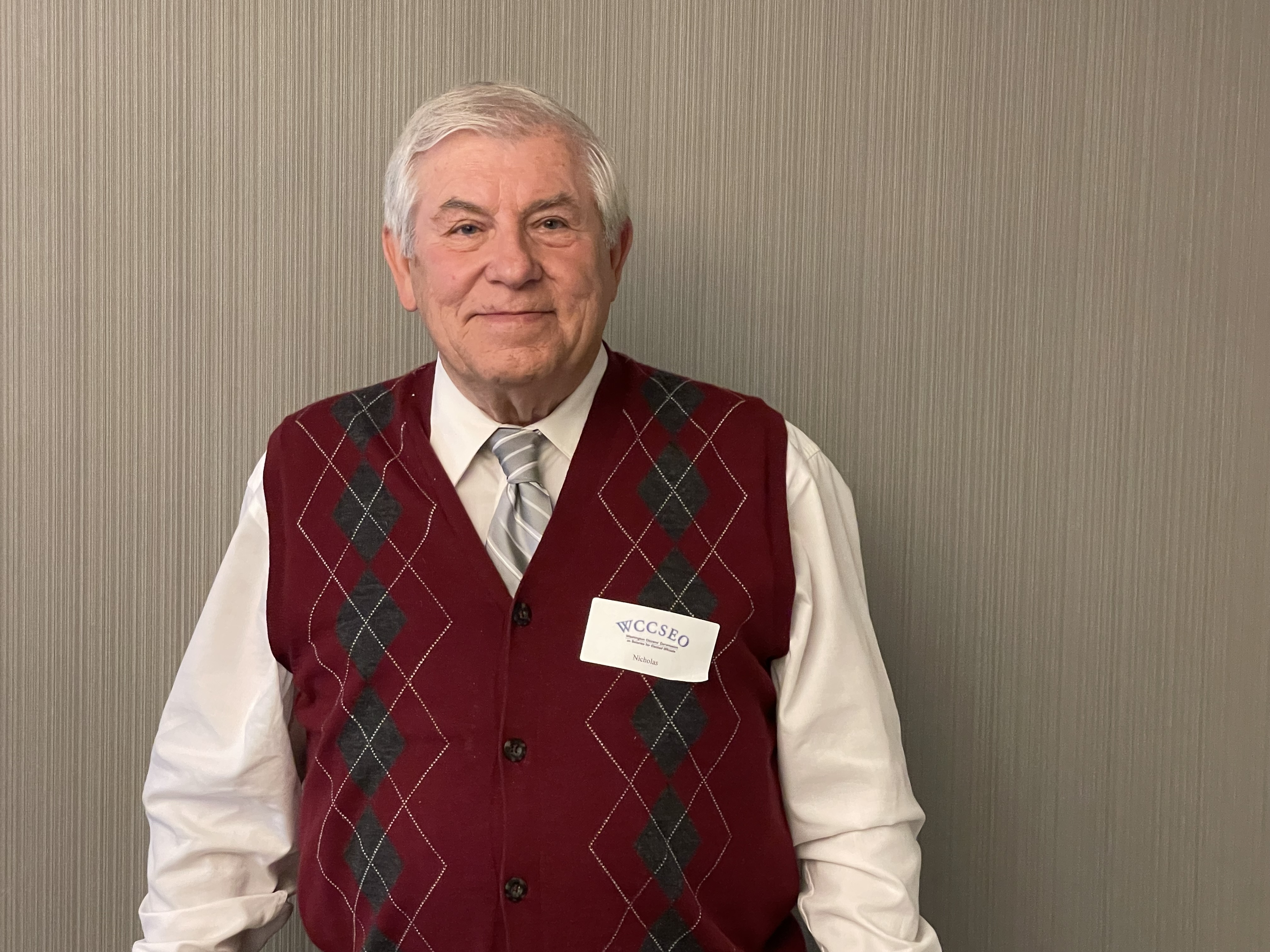 Nicholas, 80 year old man with white hair smiling wearing a white shirt, a tie and a red vest