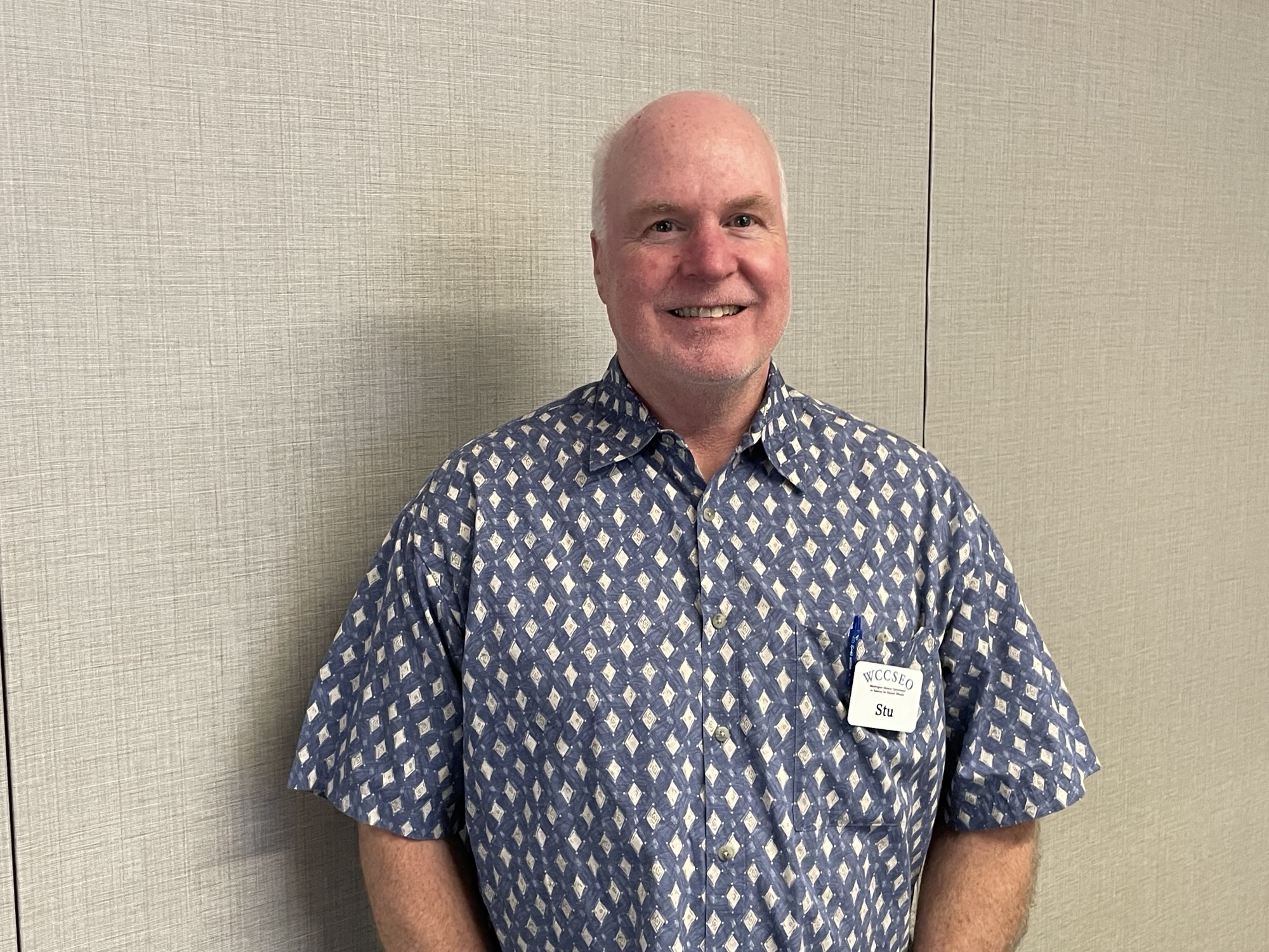 Stu, Man smiling, he has white hair and is wearing a blue button down shirt with small white diamond shapes