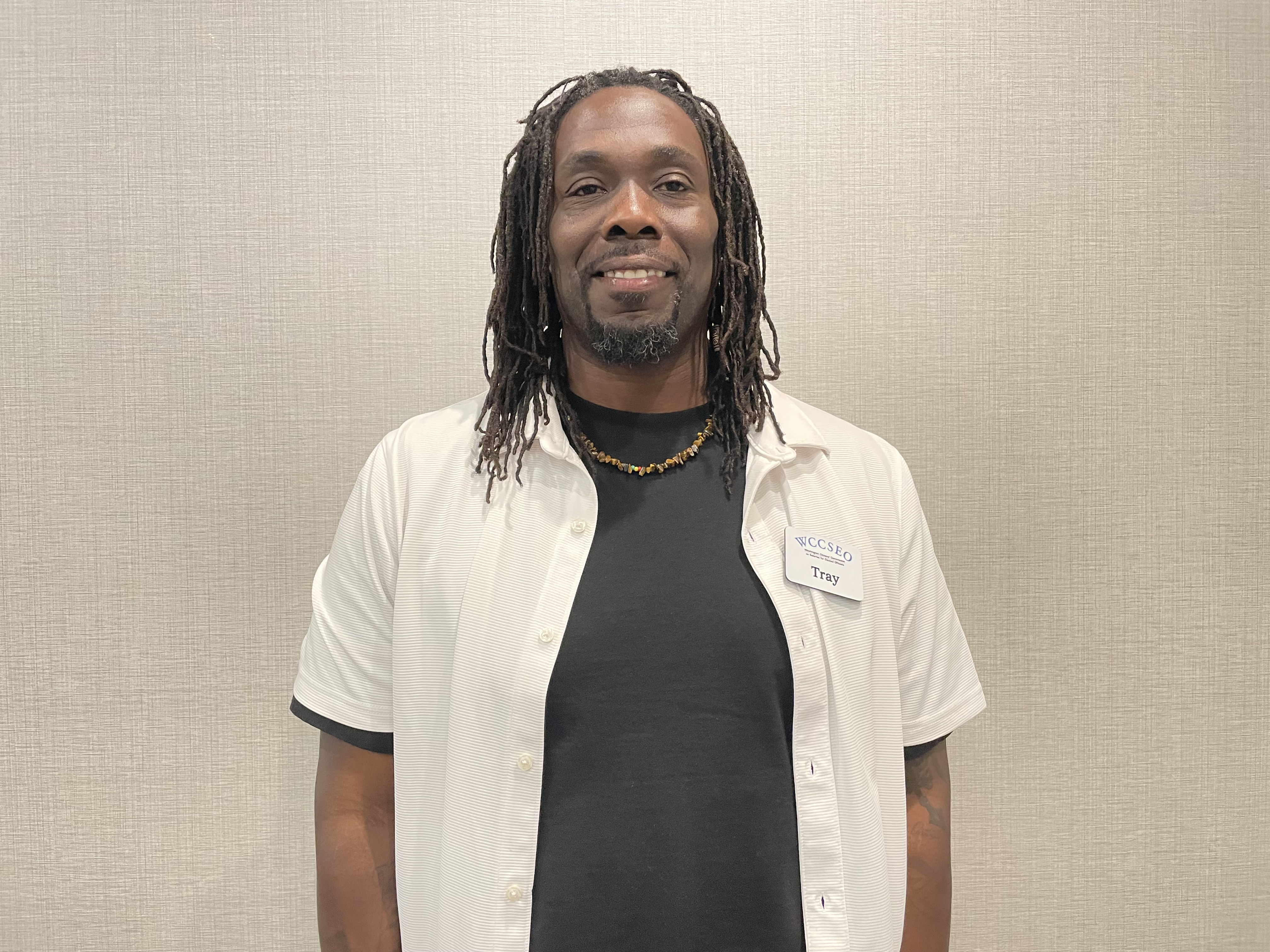 Tray, black male, shoulder length thin dreaded hair, dark complexion, with a bright smile, wearing a black under shirt and white button up